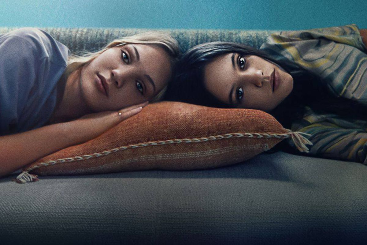 Two teenaged girls lie on the couch, their heads together.