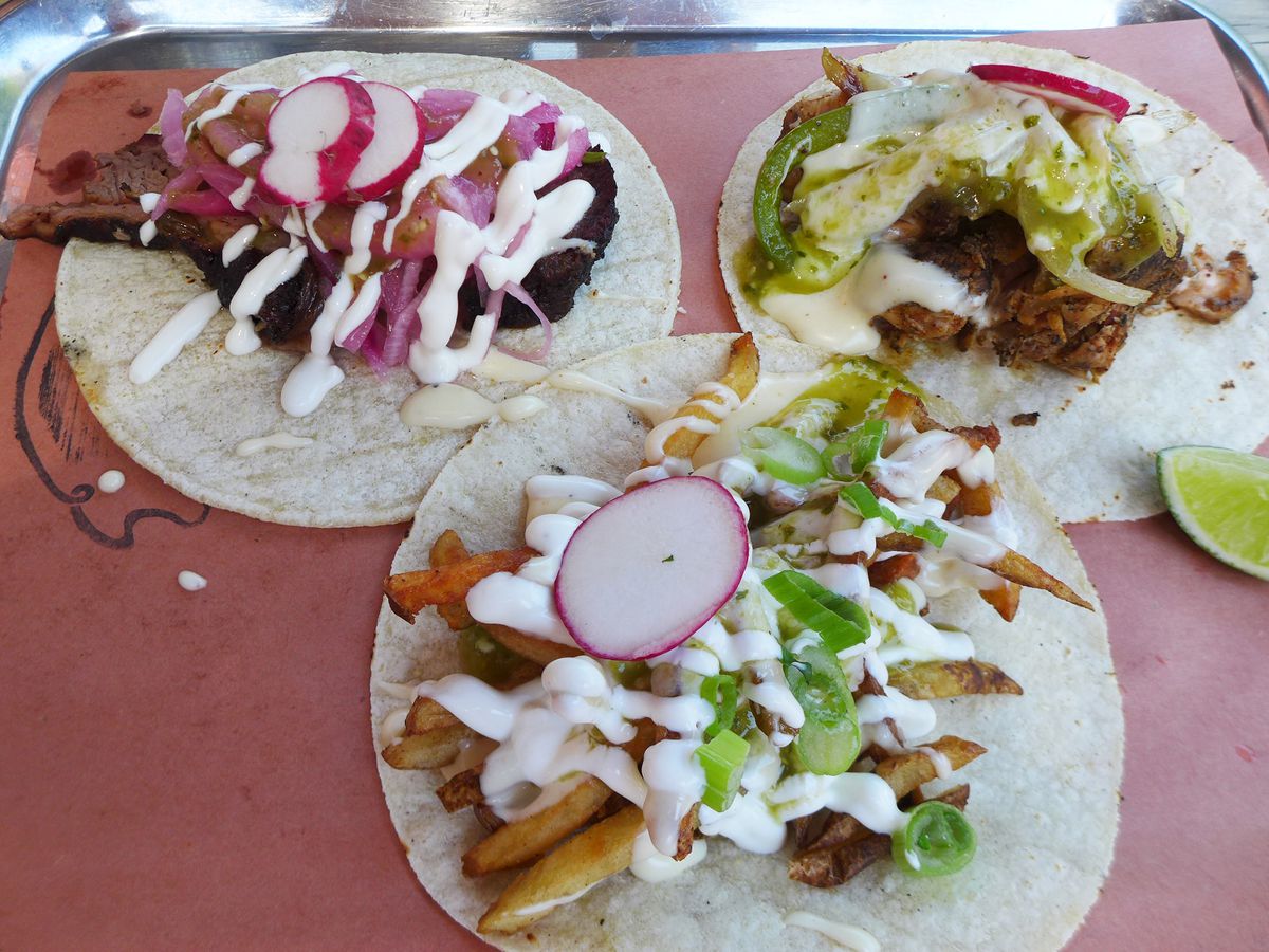 Three tacos on flattened tortillas on brown paper on a metal tray.
