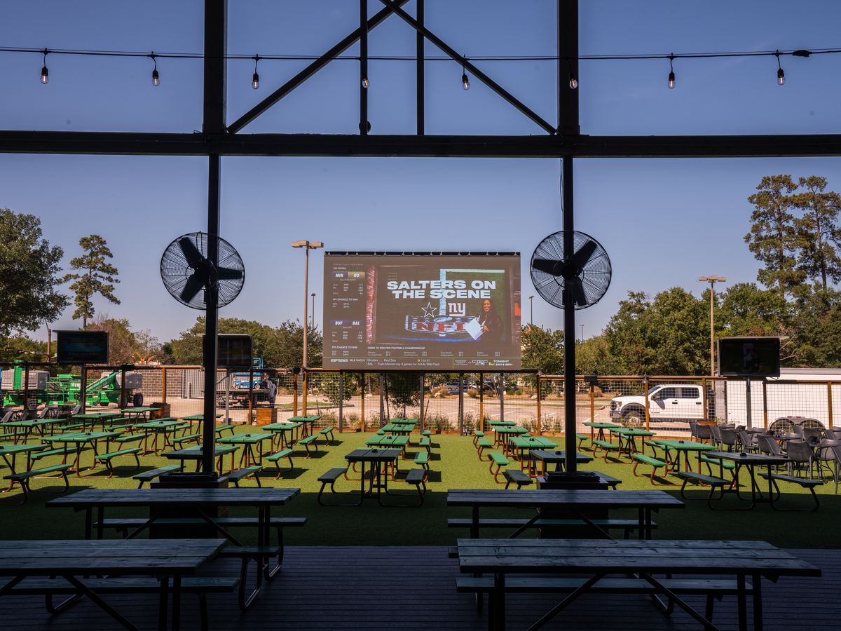 An inside view of Kirby Ice House - the Woodlands’ outdoor patio, which features a 300-inch jumbo screen, multiple picnic tables, and fans.