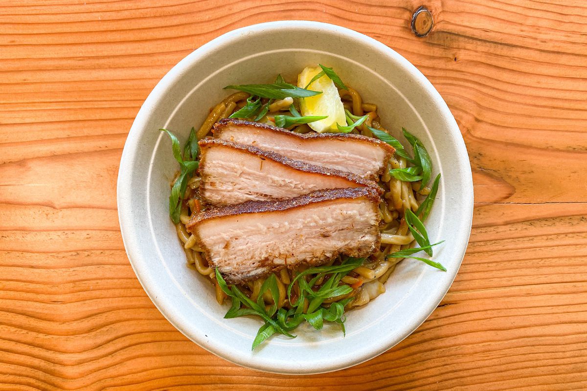 An overhead shot of a takeout bowl filled with pork belly crisped at the edges, over noodles.