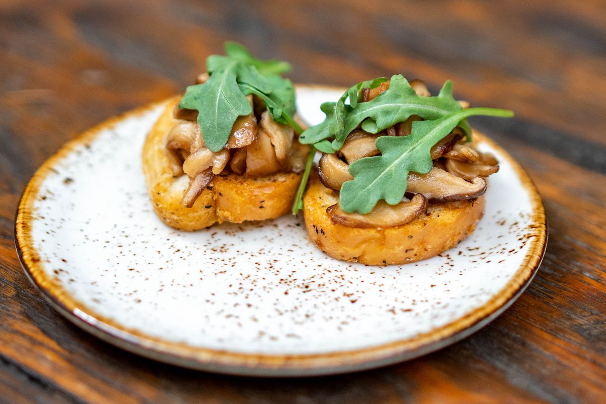 Two pieces of toasted bread on a white ceramic plate garnished with sliced ​​mushrooms and arugula leaves.