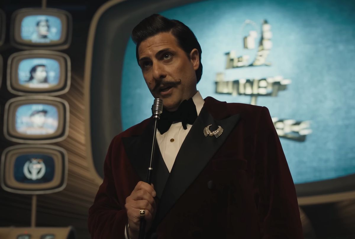 Jason Schwartzman with handlebar mustache playing Lucky Flickerman, holding a mic in front of the Hunger Games TV in The Ballad of Songbirds and Snakes