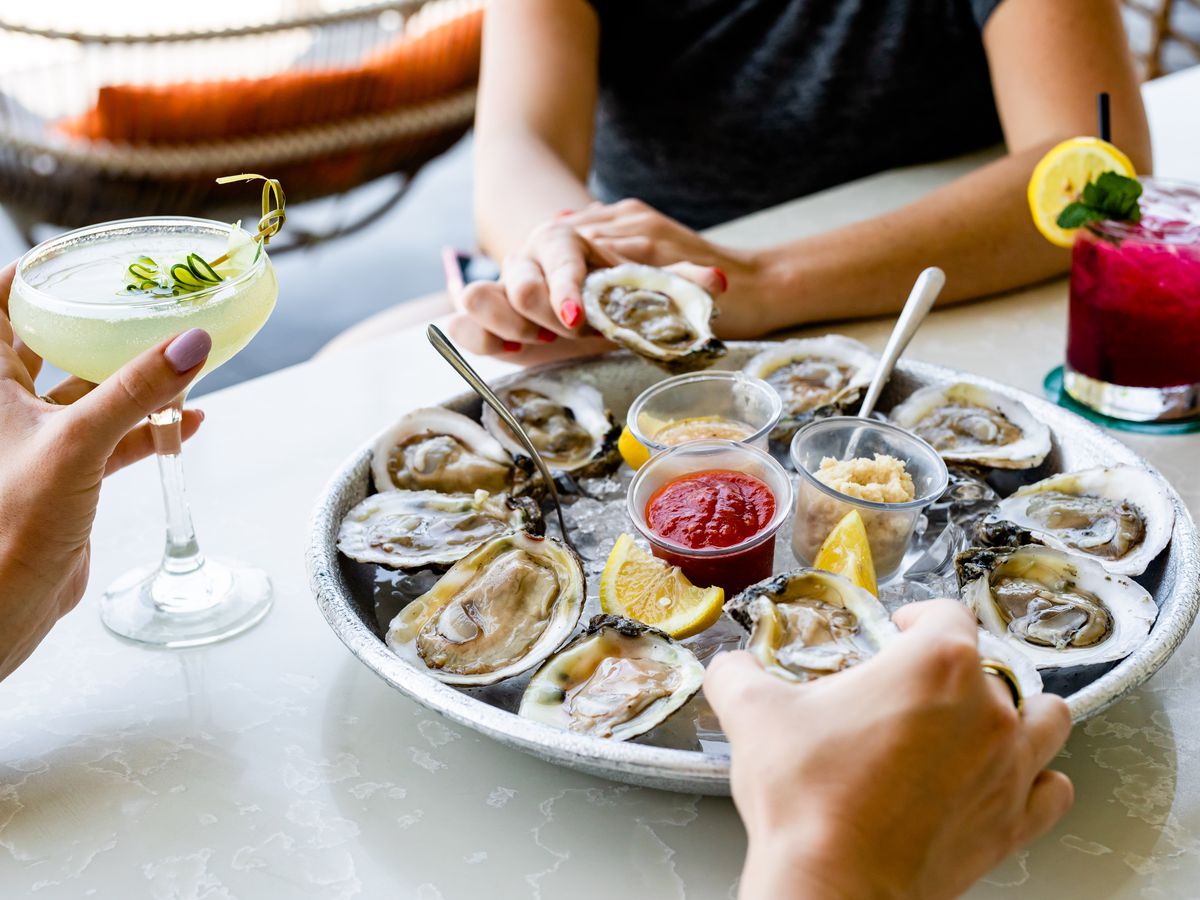 Two people picking up oysters on the half shell from a tray of ice with a dozen oysters.