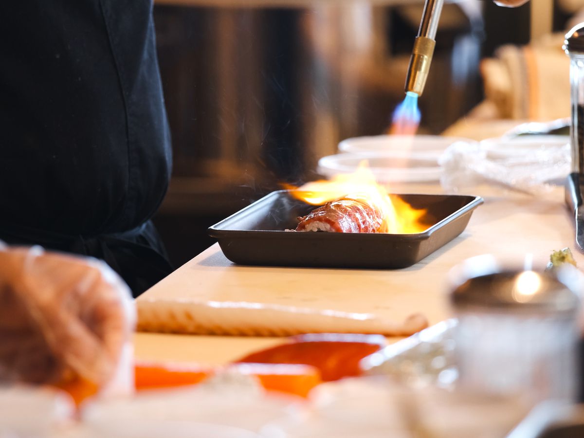 A chef with a blowtorch over a steak sushi roll. 