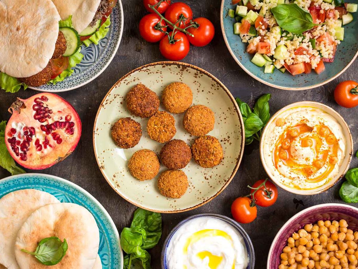 A top-down view of many dishes, including falafel, pita, hummus topped with oil and whole chickpeas, and a yogurt dip, surrounded by fresh produce like whole tomatoes on the vine and a pomegranate cut in half.