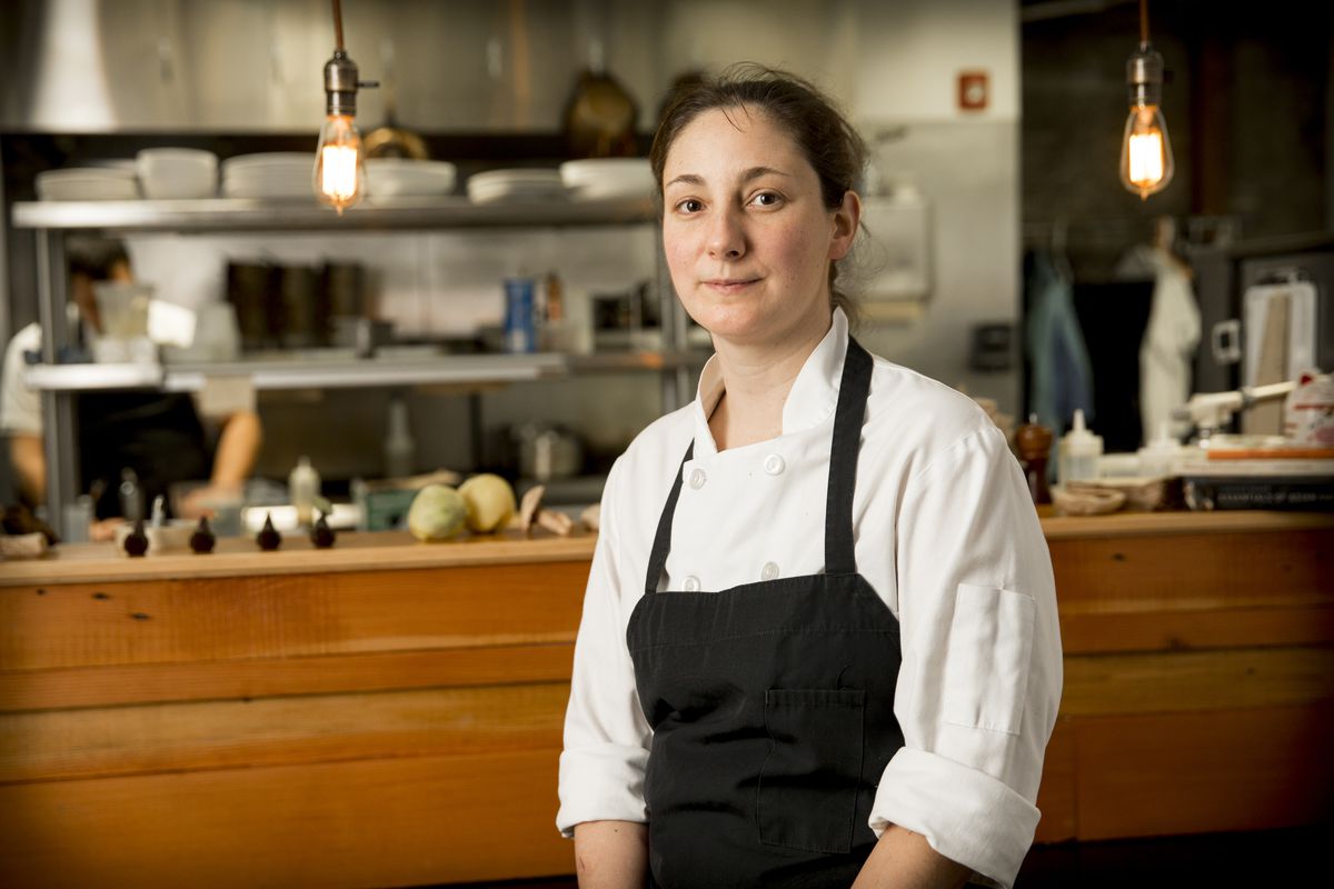 A woman in chef’s whites and a black apron sits in front of the chef’s pass at Aviary.