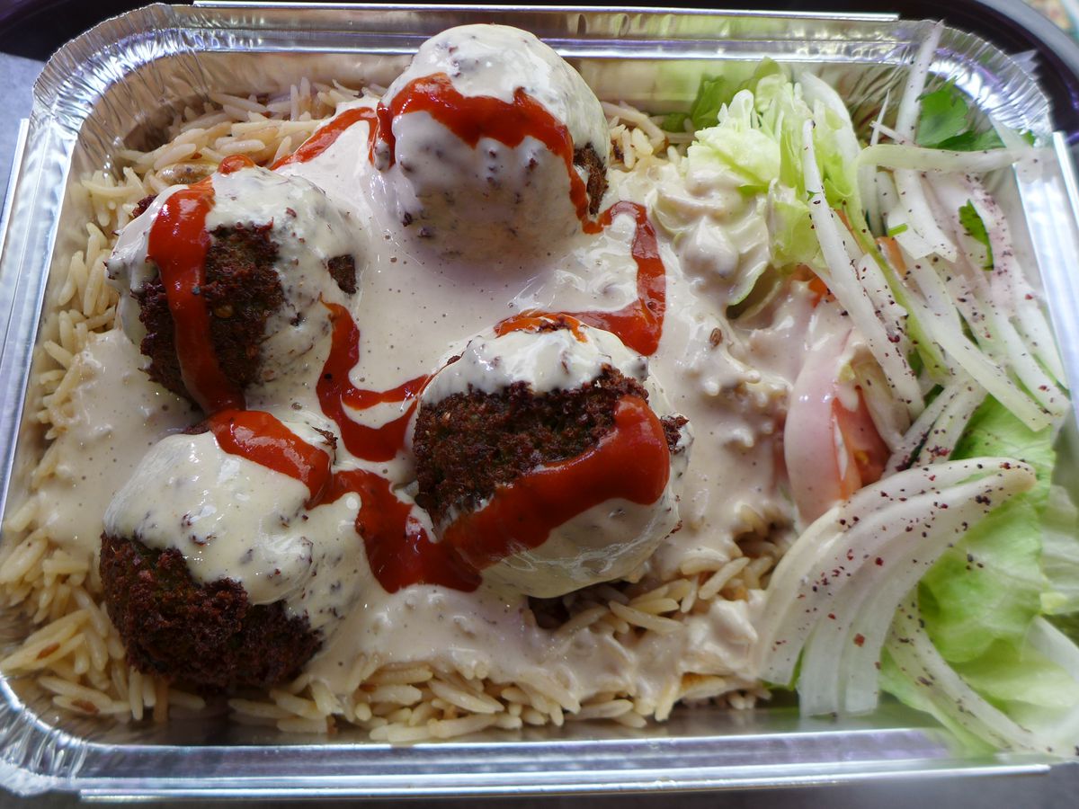 An aluminum container with falafel, lettuce, onion, and heaps of red and white sauce.