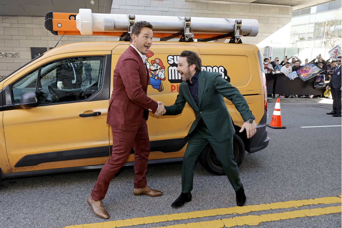 Chris Pratt and Charlie Day stand in front of a themed minivan, at The Super Mario Bros LA premiere. Pratt is in a deep red suit and Day is in a deep green suit, and the two are shacking hands with goofy expressions on their faces.