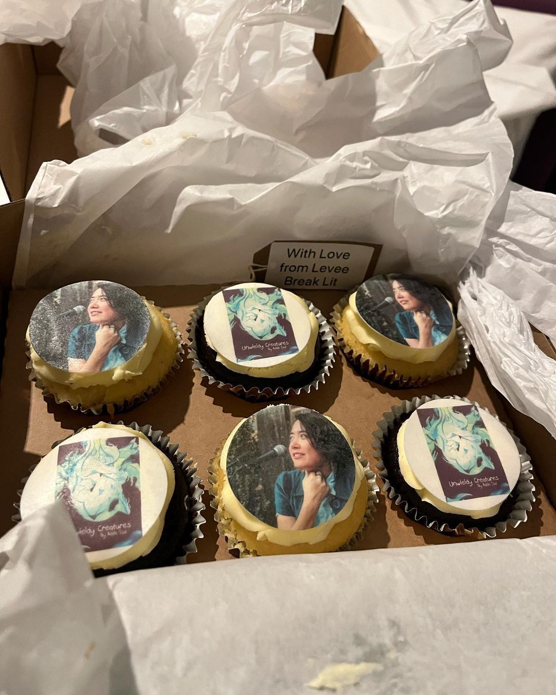 A box of six chocolate and vanilla cake cupcakes, each one topped with an edible image of either author Addie Tsai or the cover of their book Unwieldy Creatures. The cover features an abstract illustration of a curled-up person, with their head nestled into their arms, surrounded by blue and green flame-like shapes.