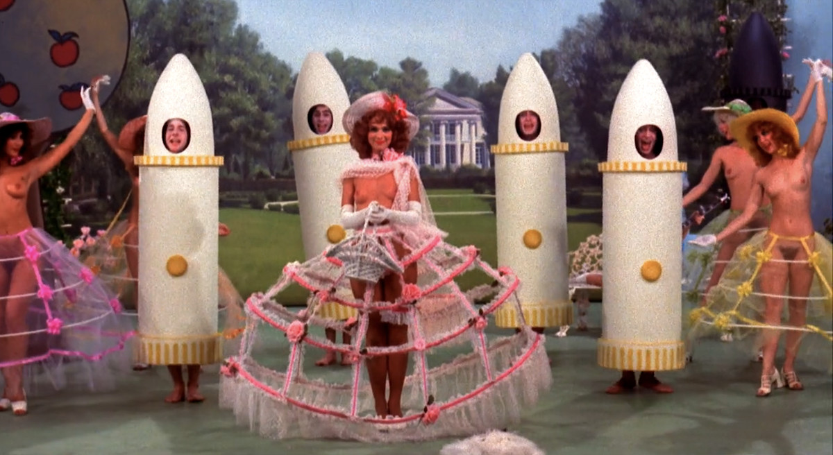 Four singing men dressed as stylized white dildos sing on stage in front of what appears to be a White House backdrop, as women wearing only Easter bonnets and translucent hoop skirt frames dance around them in The First Nudie Musical