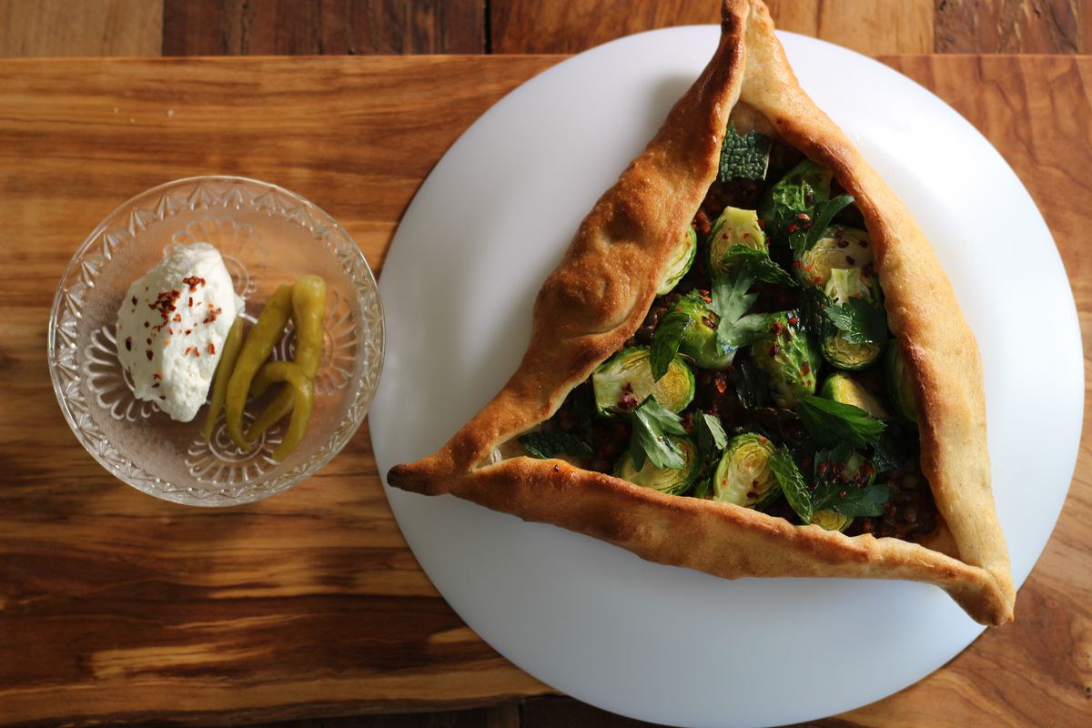Overhead view of a triangular hand pie stuffed with Brussels sprouts. On the side, there’s a small clear bowl with a scoop of a thick white sauce and a pickled garnish.
