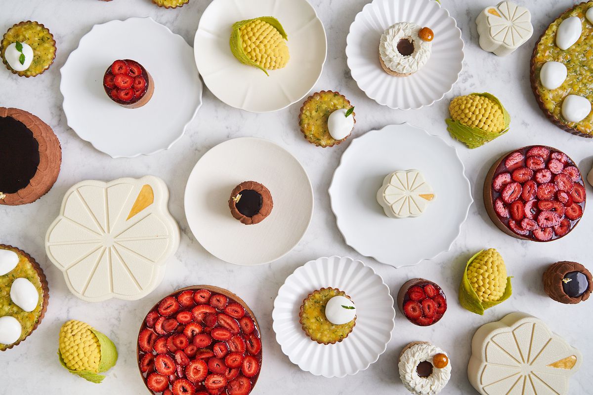 A white backdrop showcases several plates with desserts with berries, one in the shape of a lemon, and a cream-colored cake.