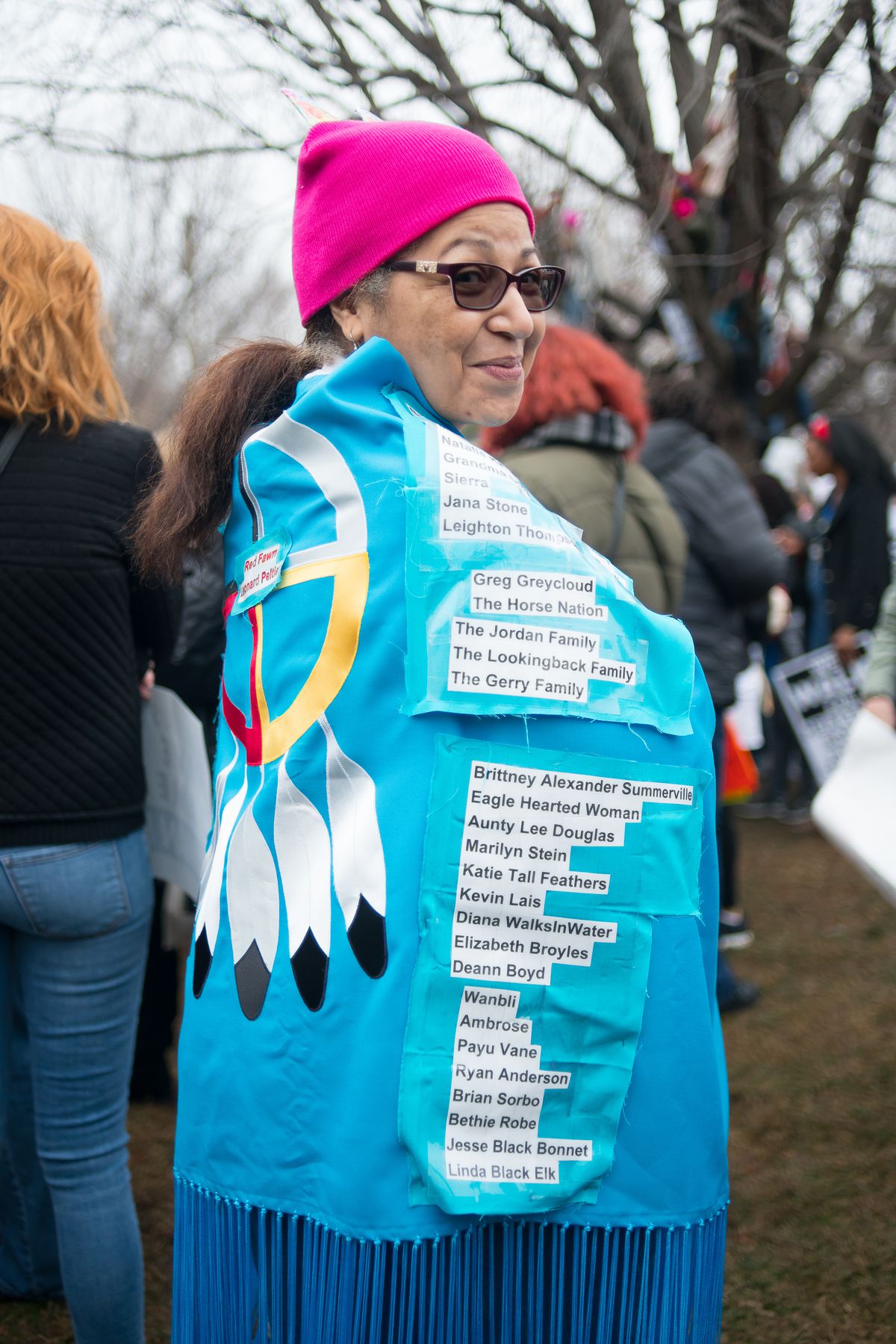 A woman smiles over her shoulder; she wears a hot pink hat, sunglasses, and a turquoise shawl with names of her friends printed out in paper and taped to it.
