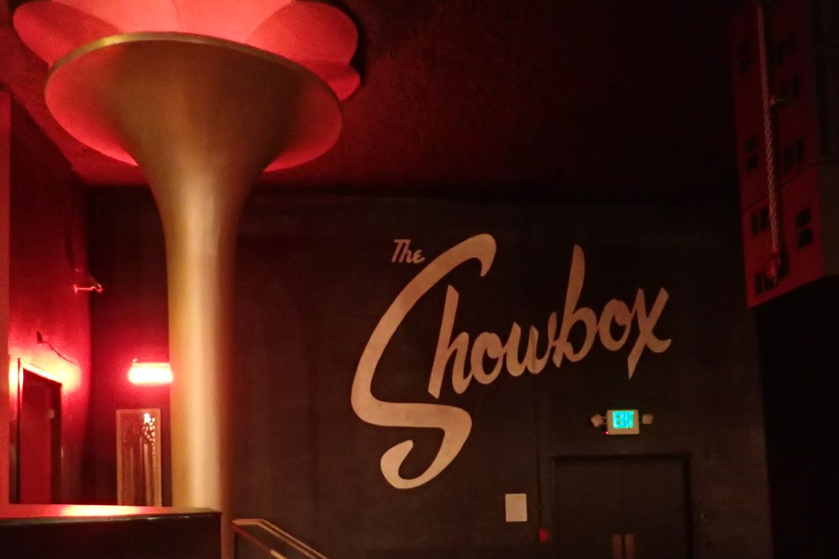 A photo shows the interior of the Showbox, including a column on the left and the venue’s logo painted in white on a black wall.