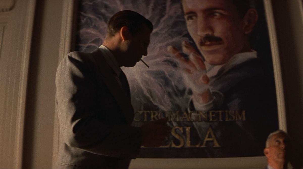 A man in a tan suit (Jeff Bridges) smokes a cigarette while walking alongside a mural of the inventor Nikola Tesla in a hallway.