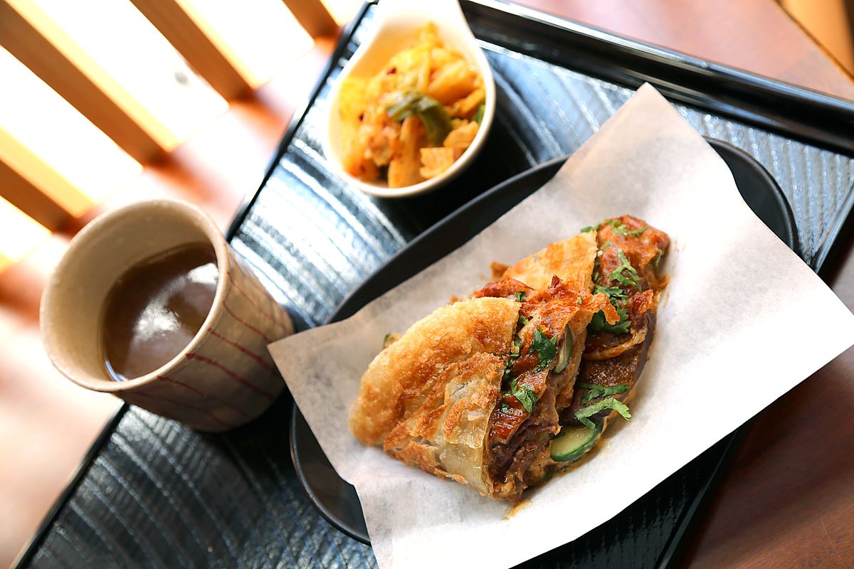 A crispy sandwich filled with meat rests on parchment paper in a plastic dining tray. Beside it, there is a dark beverage and a side of pickled vegetables.