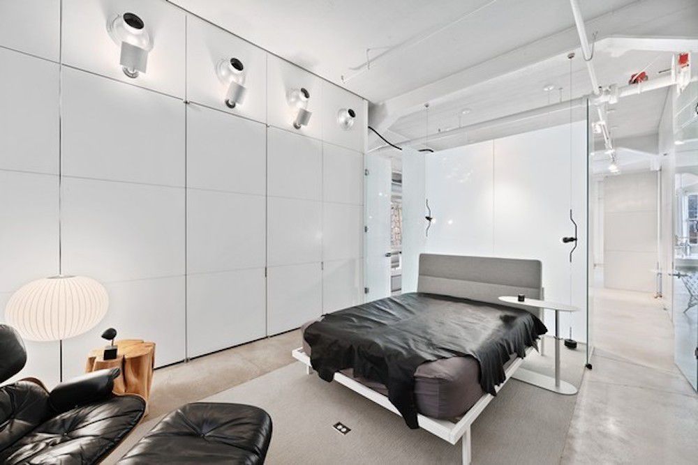 A contemporary bedroom with white walls, a gray bed, and a small seating area. 