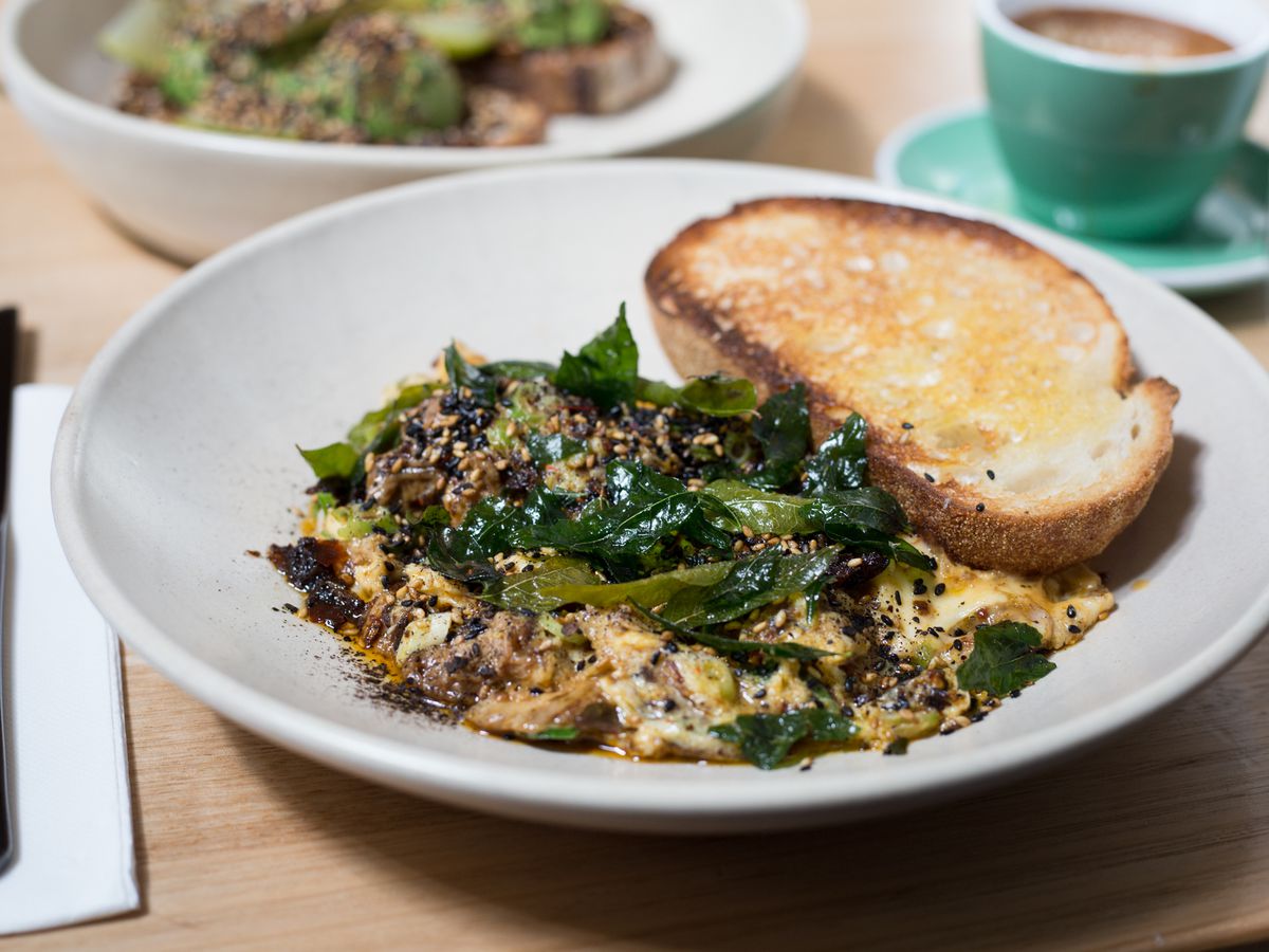 A shallow bowl full of eggs scrambled with duck meat, studded with curry leaves and lemongrass, with a thick slice of sourdough on the side, with other dishes blurred in the background. 