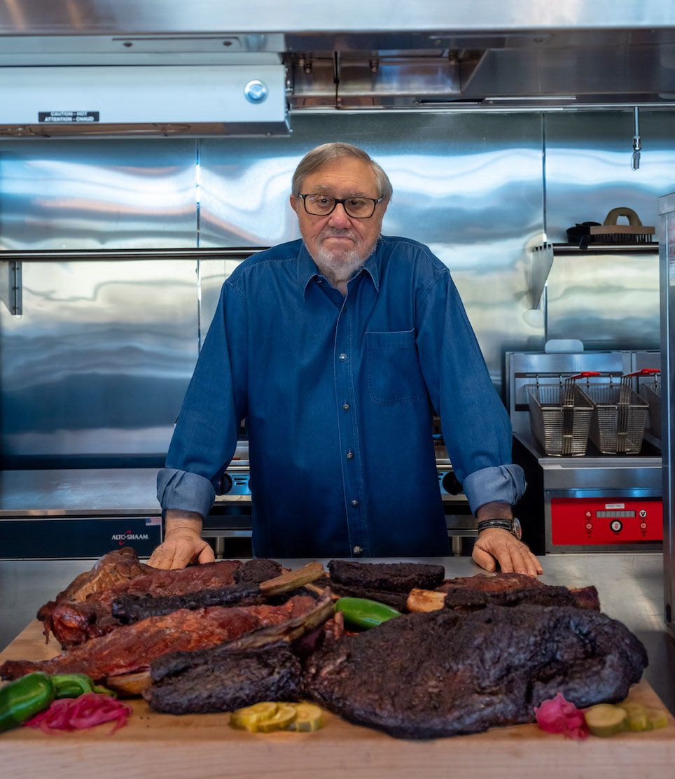 An older man stands behind a kitchen counter. In front of him is a mound of barbecued meats.