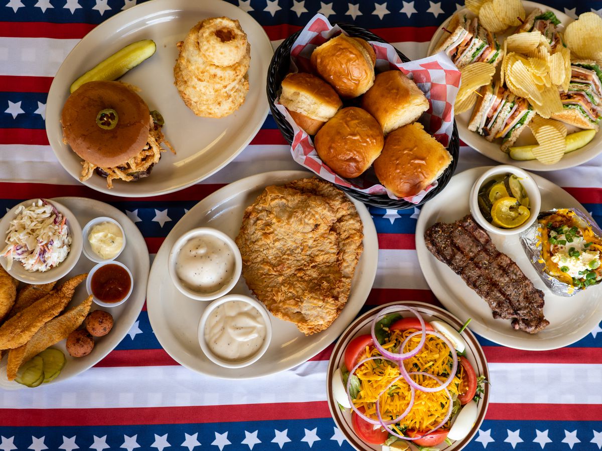 A table with a flag tablecloth holds multiple dishes on oblong, off-white plates, including a club sandwich with chips, a strip steak, a salad, a chicken fried steak with mashed potatoes and squash, fried catfish, and a burger.
