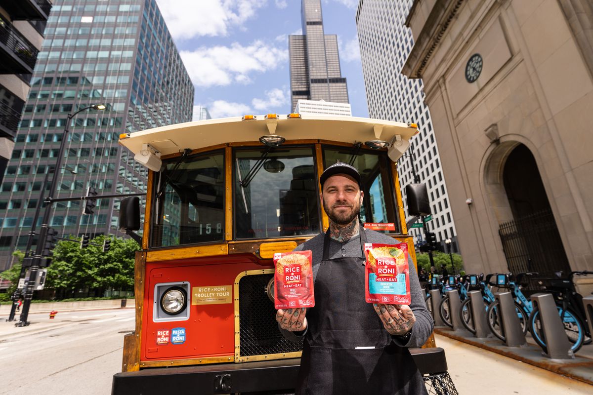 A heavily tattooed male chef poses in front of a trolley car holding two boxes of Rice-A-Roni.