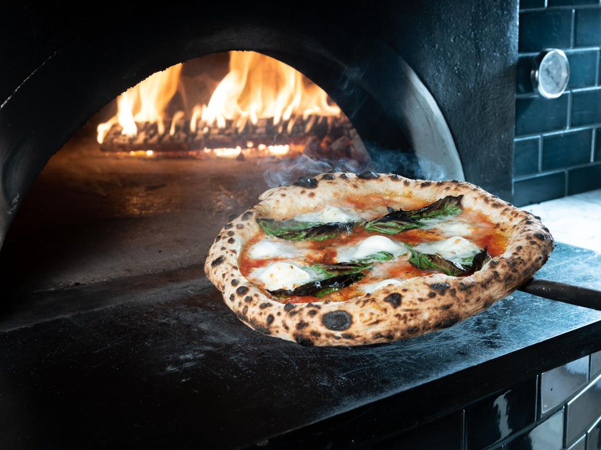 A blistered pizza is pictured next to the mouth of the restaurant’s wood-fired pizza oven, with flames visible near the back of the oven.