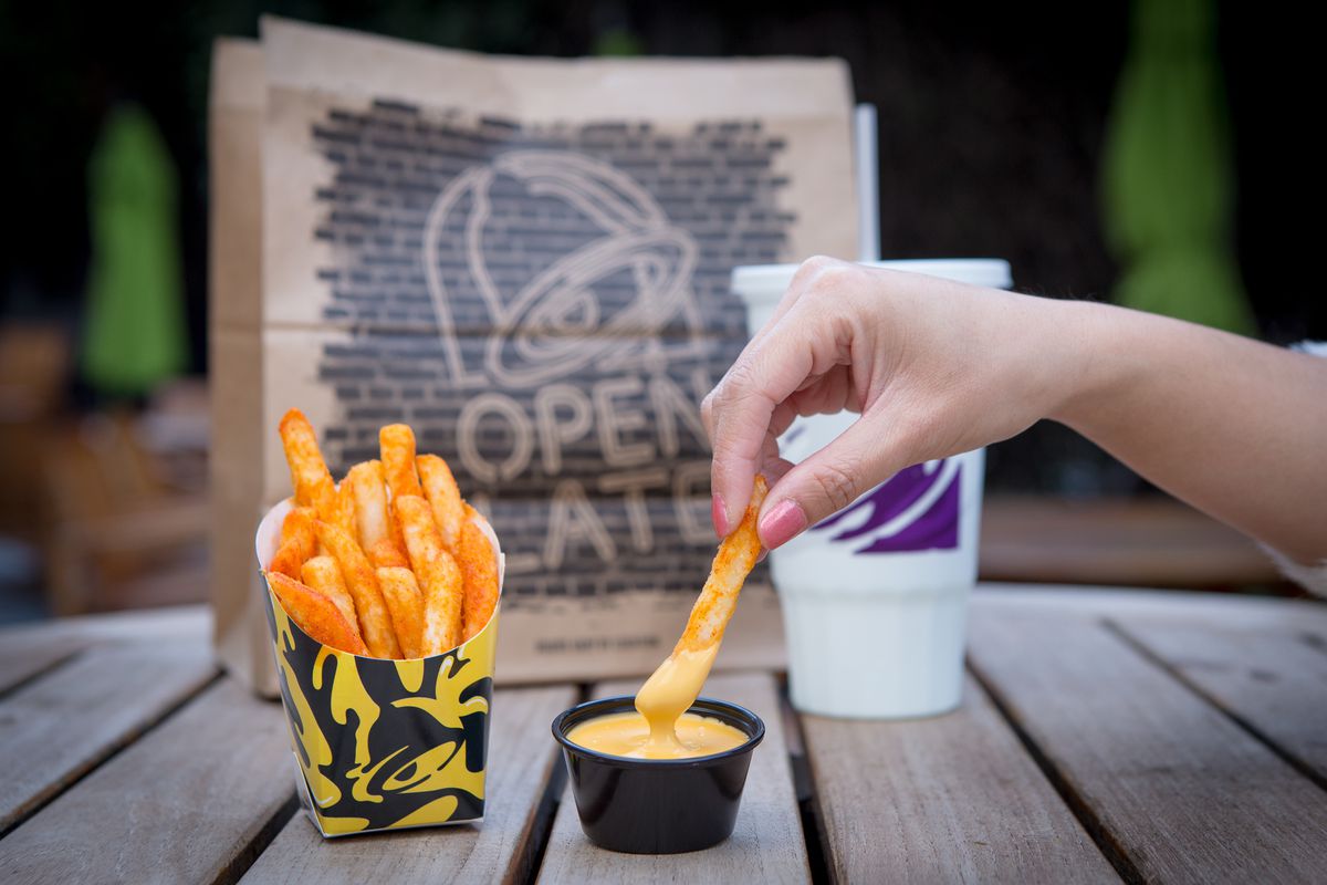 A container of Taco Bell nacho fries sitting next to a black plastic ramekin of nacho cheese. A white person’s hand is dipping a fry into the cheese.