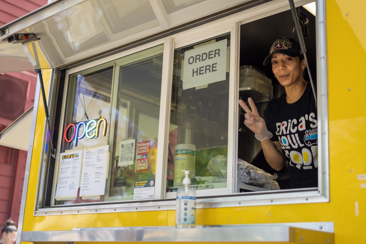 A woman in a hat poses with a peace sign standing in a yellow food cart window, wearing a shirt that says “Erica’s Soul Food.”