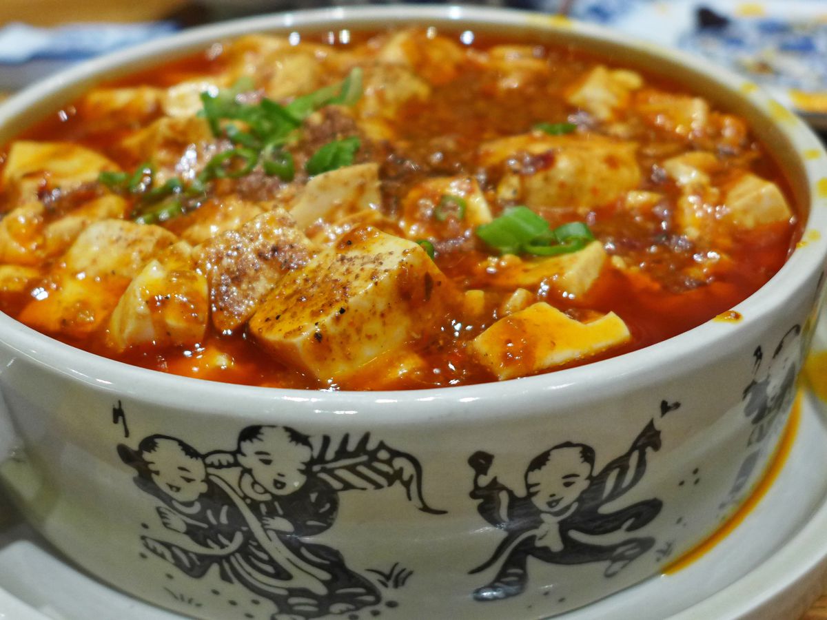 A white bowl with blue children depicted on the sides filled with white bean curd in red oily sauce.