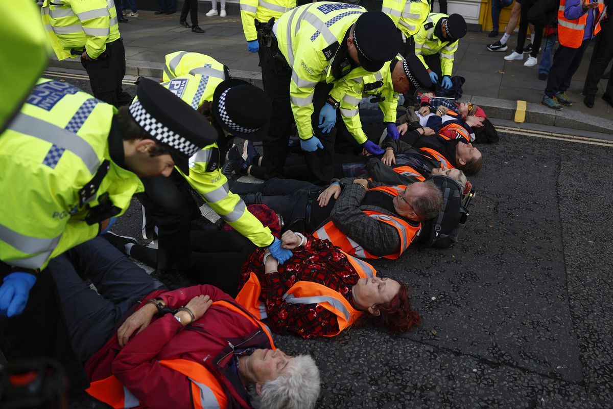 Middle aged protesters, in neon orange vests, lie on their backs in the middle of a street. They are being handcuffed by a gaggle of officers in neon yellow jackets.