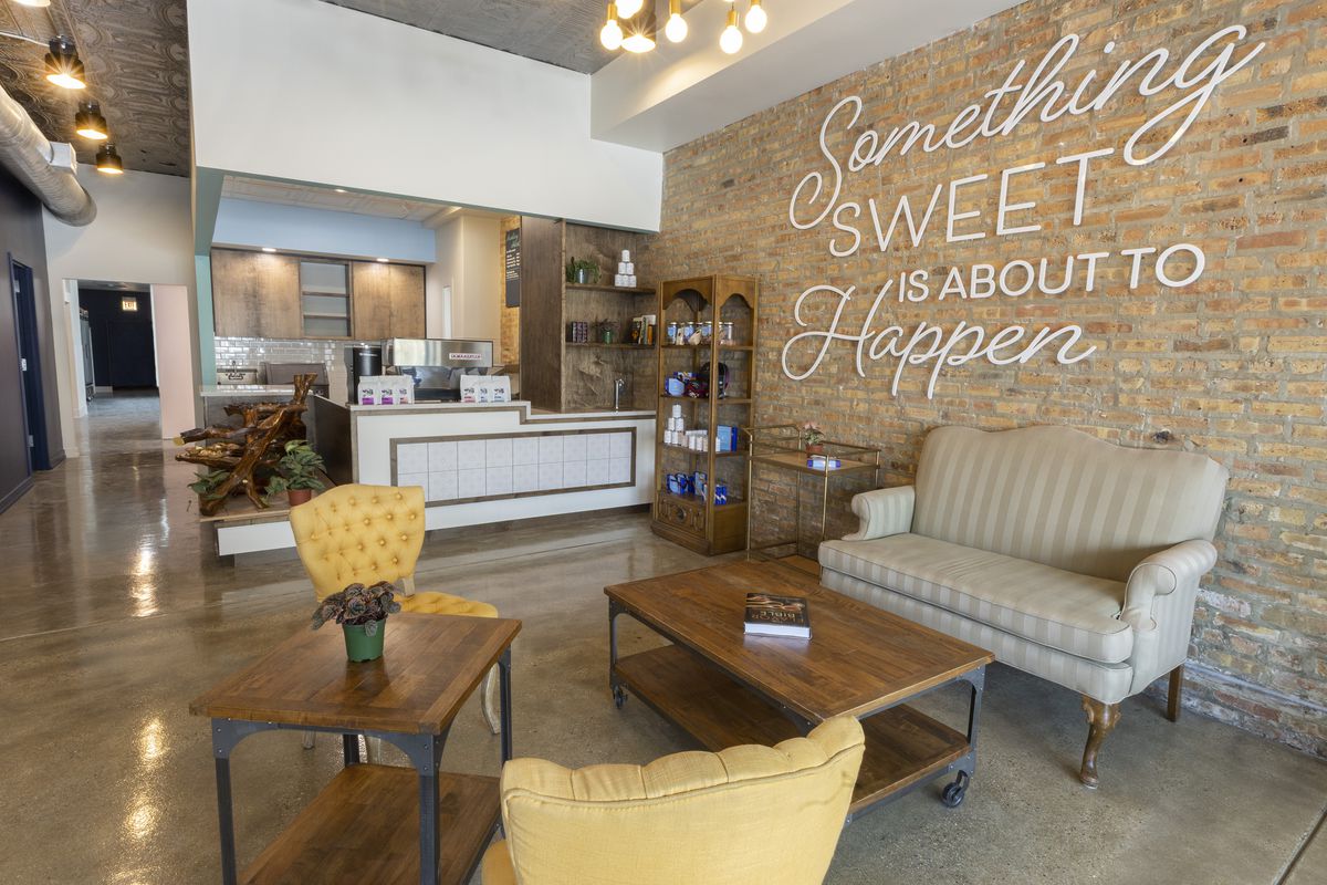 An airy cafe space with exposed brick walls and a wall display that reads “Something sweet is about to happen.”