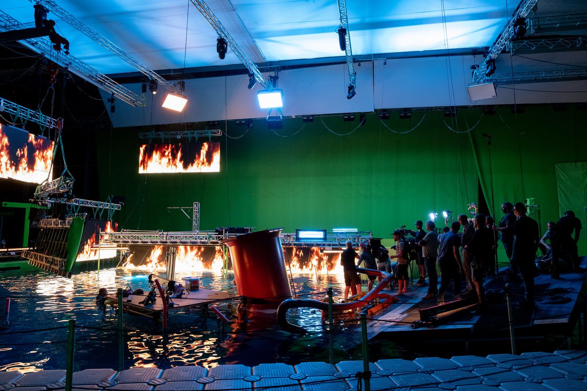 The wreckage of a ship built on a stage with screens projecting photoreal flames and a green screen backdrop. James Cameron stands on a platform with his crew directing an actor during the making of Avatar: The Way of Water.