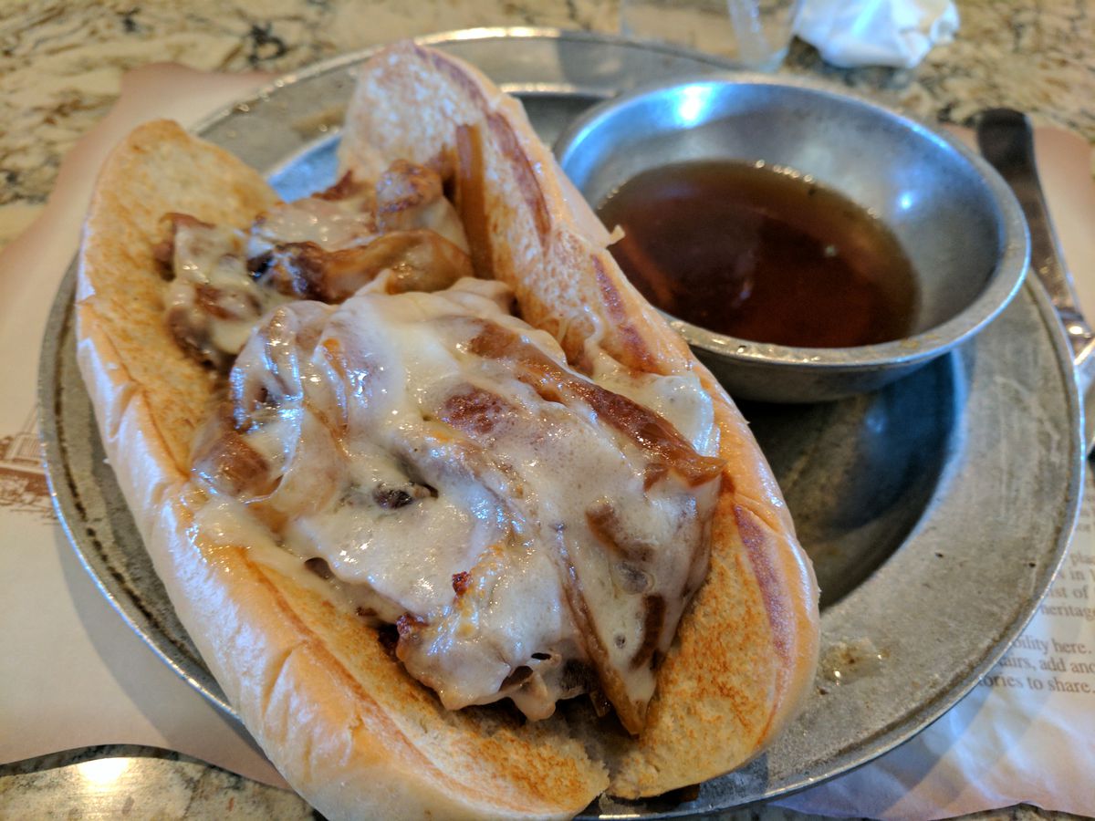A cheese-covered French Dip sandwich on a pewter plate with au jus on the side.