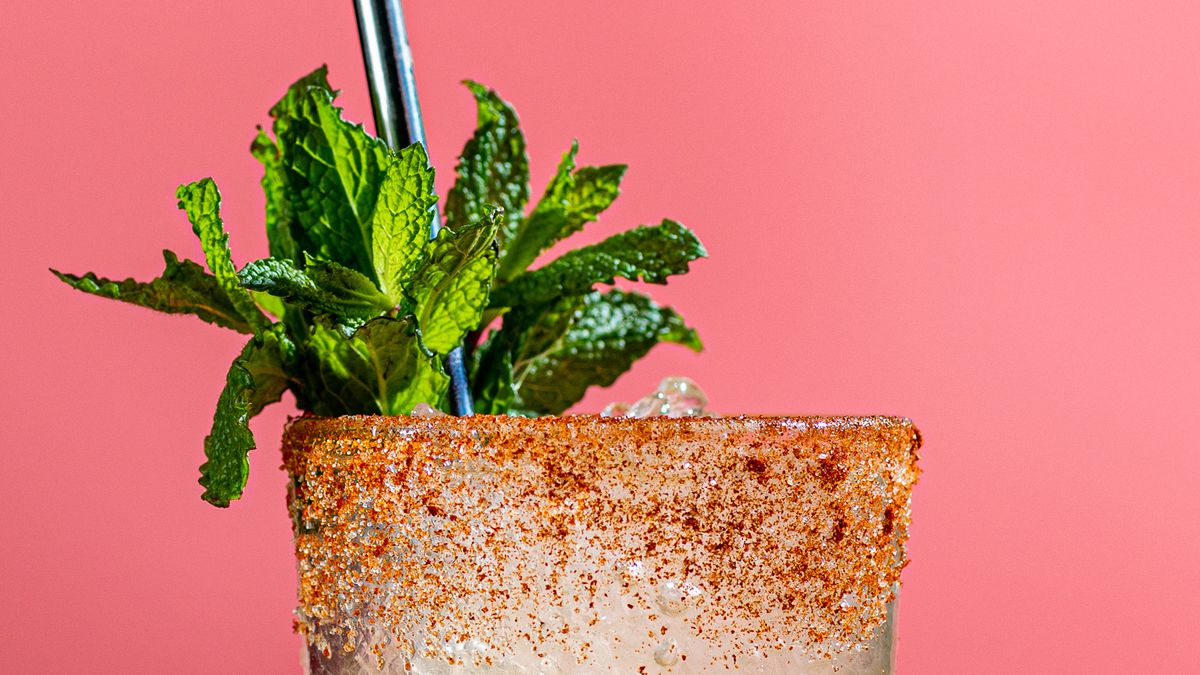 A yellow cocktail drink with a sprig of mint against a pink background.