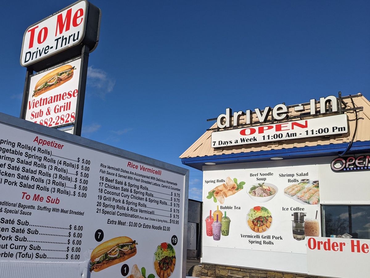 A small drive-thru exterior, with a menu with images alongside a longer print menu. A large sign for To Me Vietnamese Sandwiches hangs overhead in the background