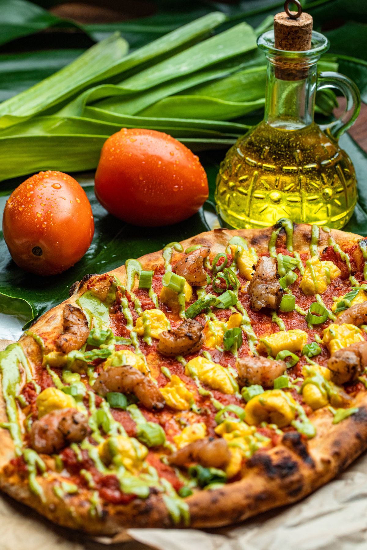 A close-up photograph of a flatbread pizza topped with meat, sauce, and green vegetables. In the background, two tomatoes sit next to a bottle of oil.