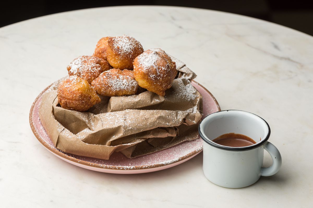 A pile of fried dough balls on a pink plate resting on paper bag with side of chocolate in a cup.