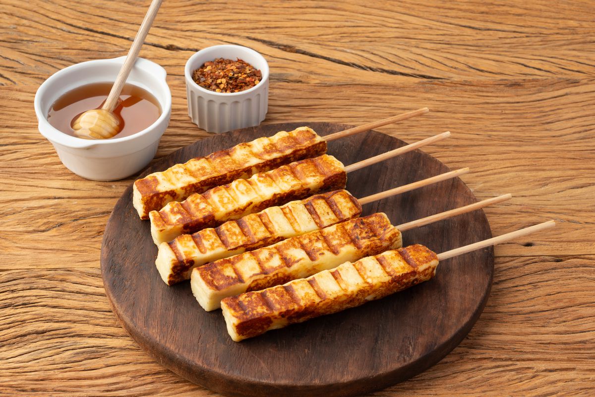 Five skewers containing a white spongy cheese and grill marks on the exterior sit on a plate. Honey and pepper flakes sit in ramekins alongside.