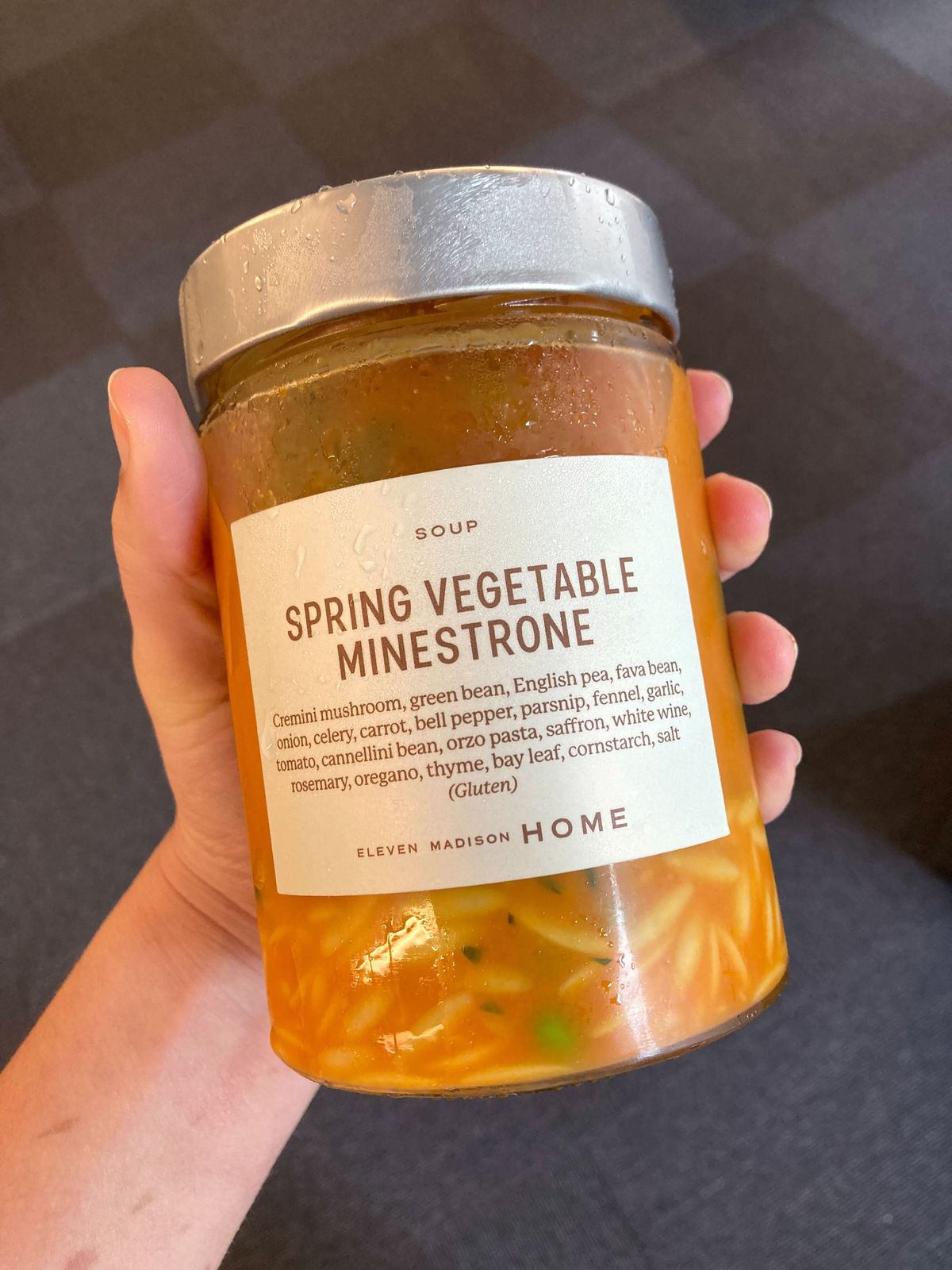 Hand holding up glass jar containing orange liquid and noodles; label on the jar reads “Spring Minestrone Soup” along with an ingredients list.