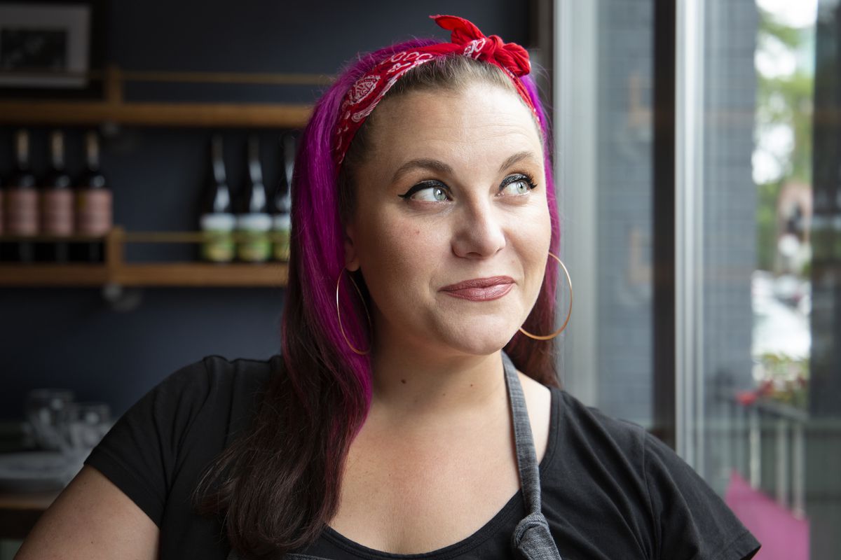 Closeup portrait of a woman with a slight smile, looking toward a sunny window to the right. Her hair is bright pink and secured with a red bandana headband. She stands in front of shelves of wine in a restaurant and wears an apron.