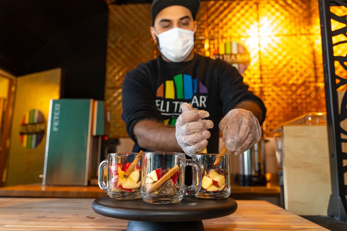 A man in a mask and plastic gloves prepares three drinks in glass mugs with apple slices and cinnamon sticks