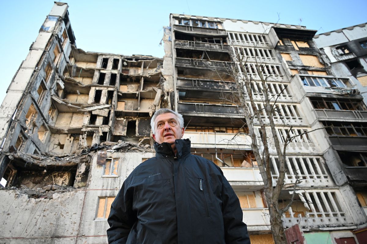 A man stands before a destroyed apartment building, some of which has caved in, some of which is burned-out.