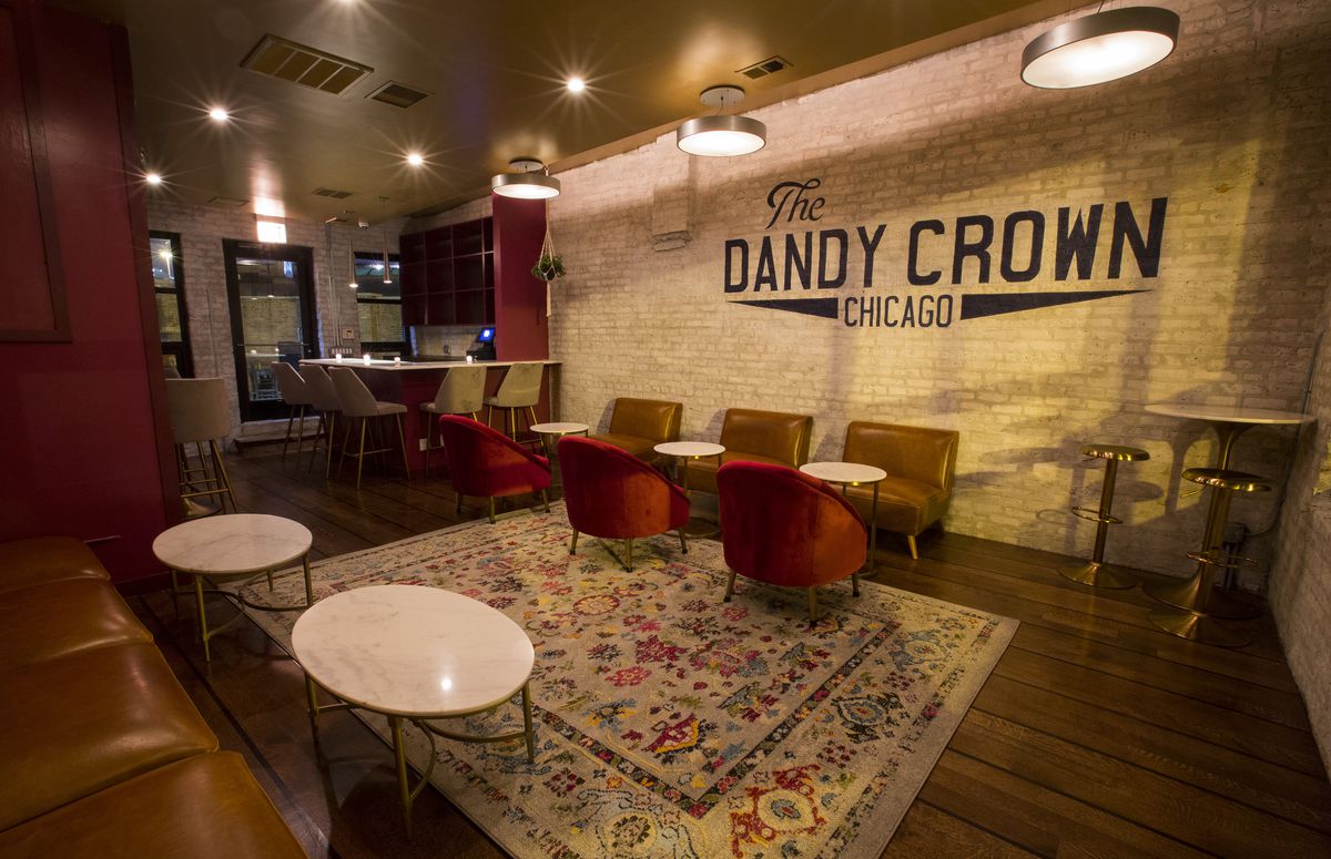 A large drinking space with white painted brick walls that read “The Dandy Crown Chicago” with red furniture.