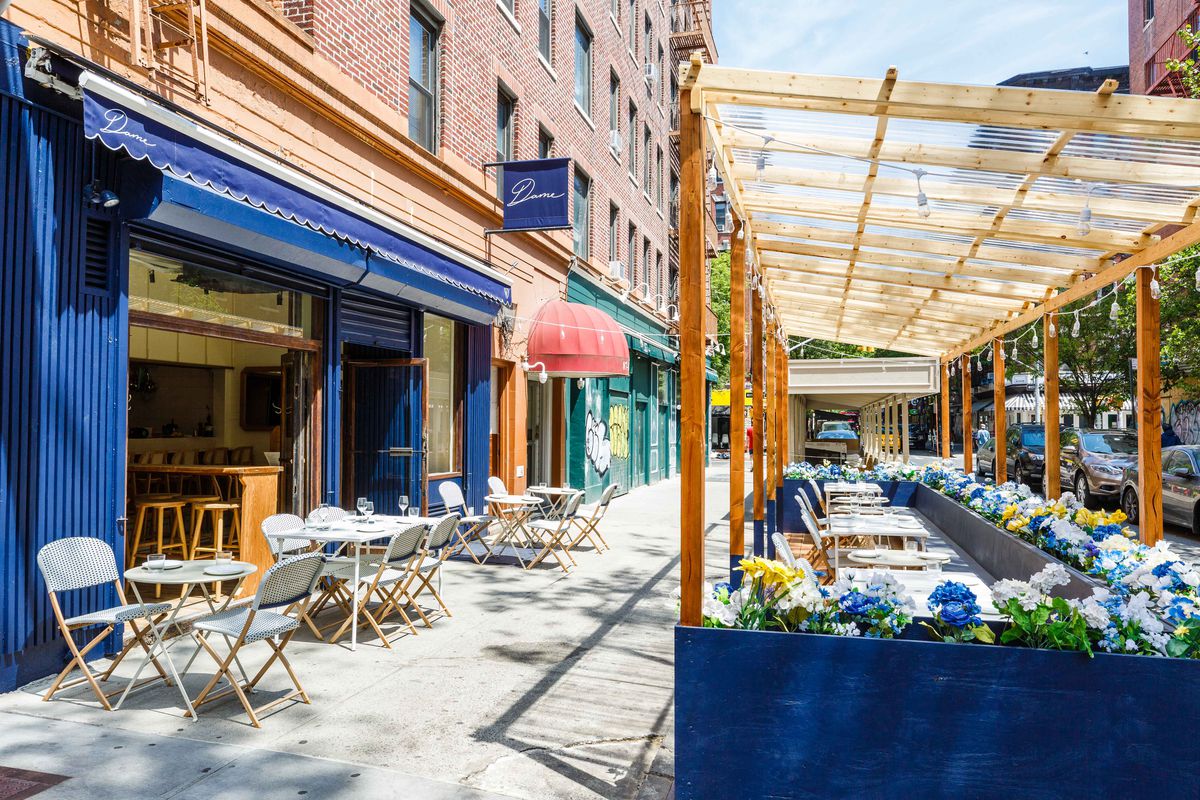 A blue restaurant facade with white tables and chairs on the sidewalk out front. A painted plywood patio sits curbside on the sidewalk with blue, white, and yellow flowers planted around the edges.
