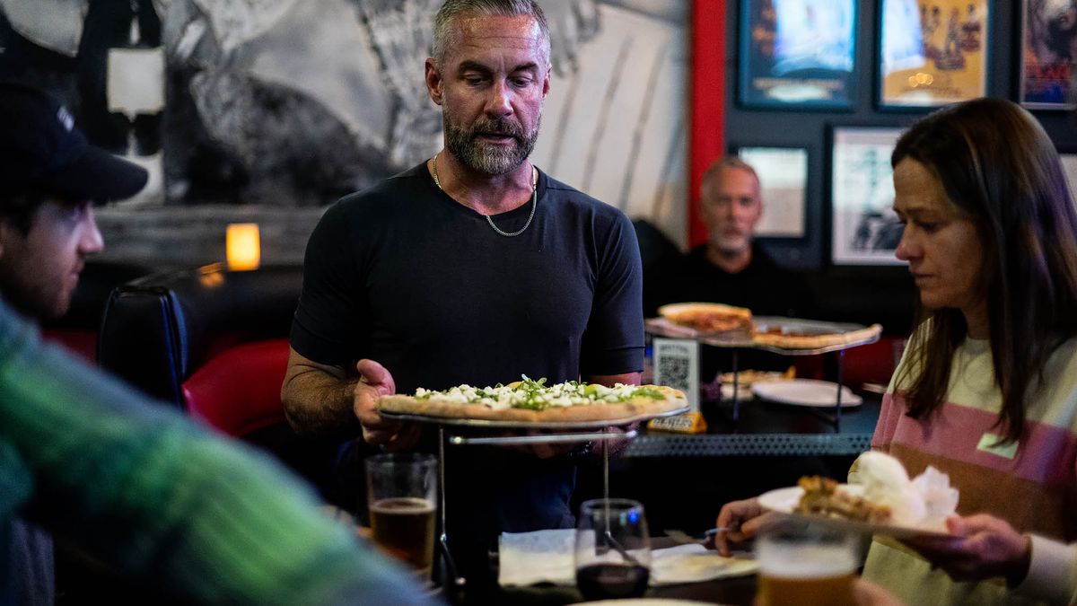 A man with silver hair and a dark t-shirt places a pizza on a table at a dark restaurant.