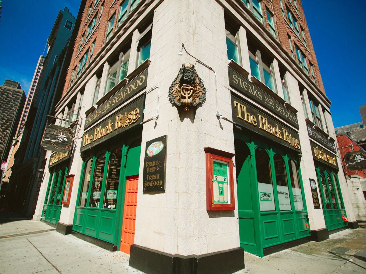 Exterior view of a bar, with the light stone corner of the building centered in view. Both the left and the right sides of the bar have a green exterior and black signage with the Black Rose written in gold