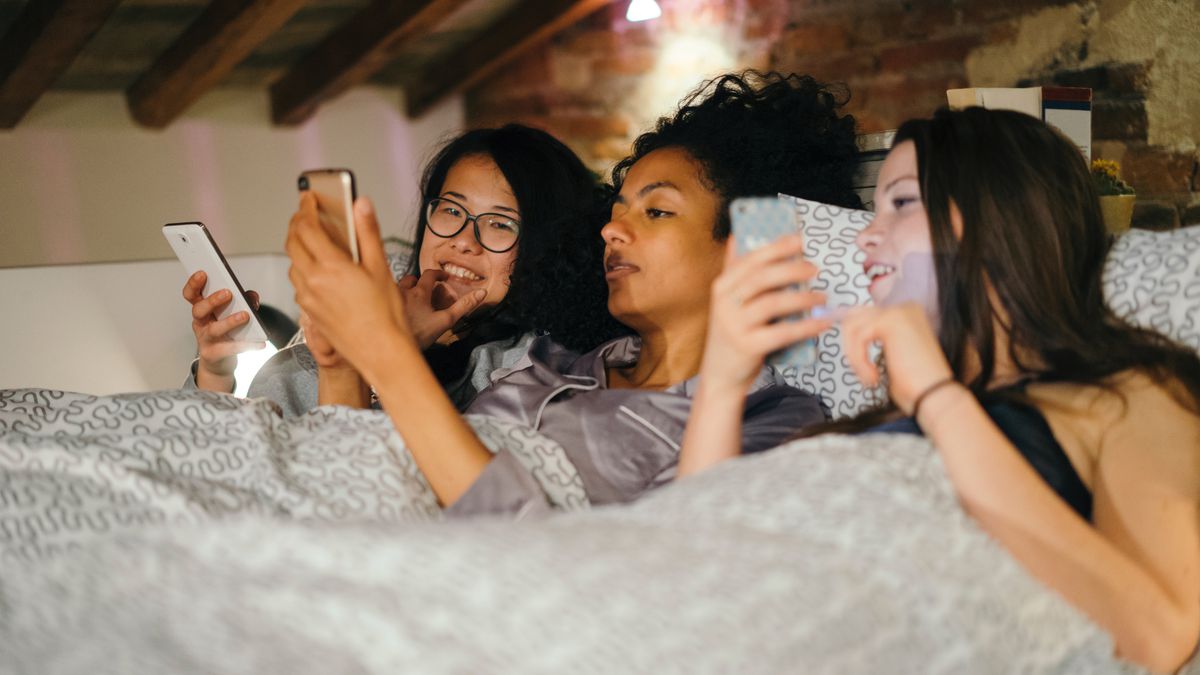three women lie in one bed using their phones