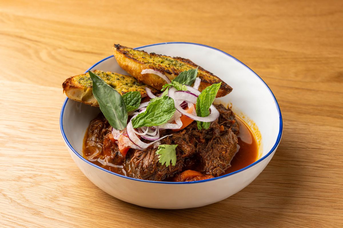 A bowl of braised red meat in a white bowl on a wooden table with a blue lip.