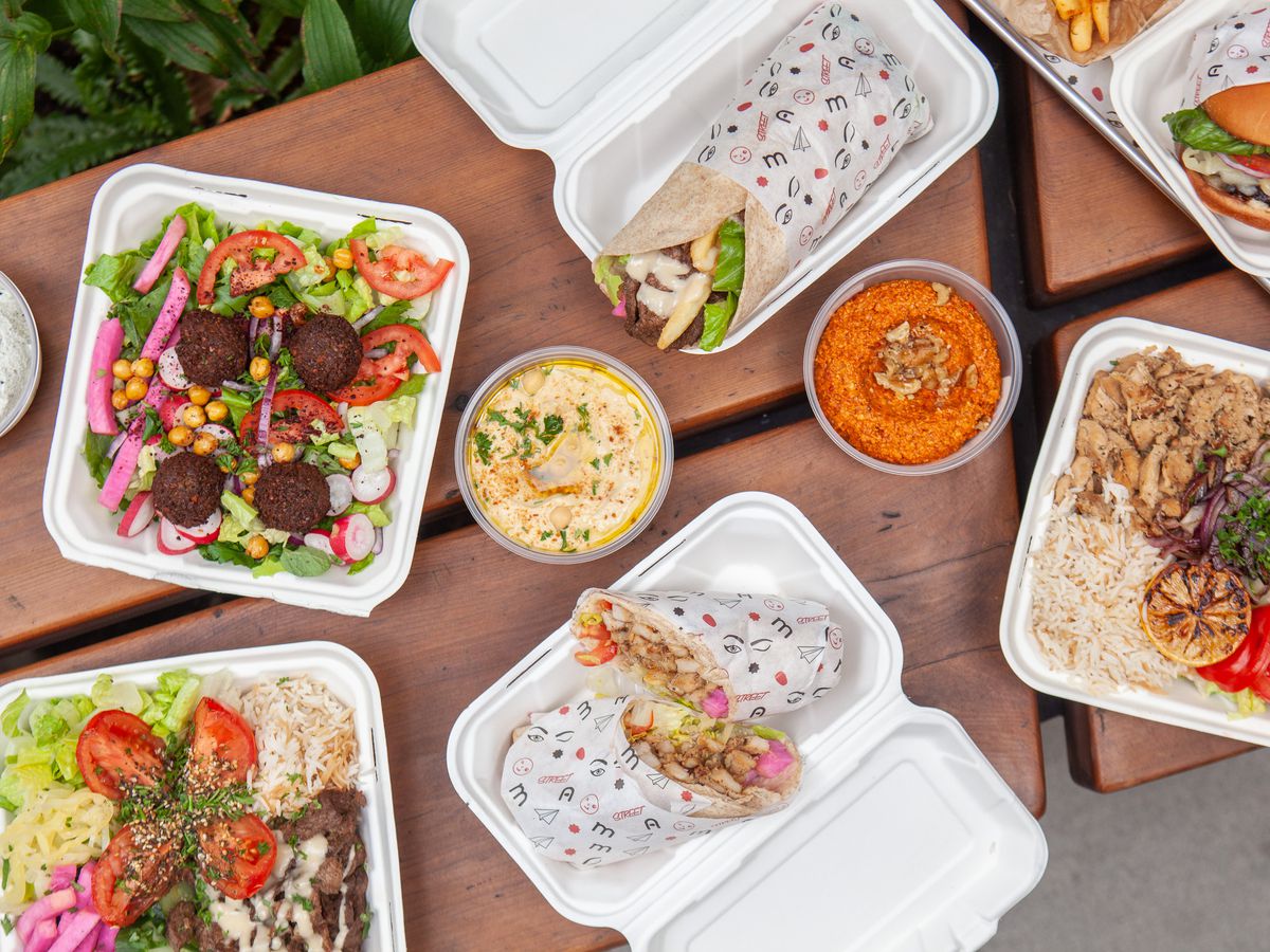 A spread of dishes in white cardboard takeout containers on a wooden picnic table: chicken on rice, falafel salad, wraps, sandwiches, and more.