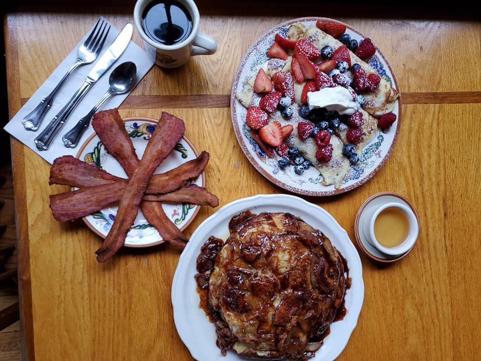 Topdown view of three plates: the top left one has bacon, the top right one has berry-covered crepes coated in sugar, and the bottom dish is a pile of pancakes covered in nuts and syrup